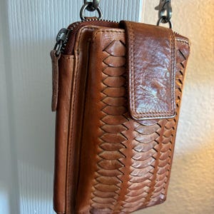 Puede incluir: Un bolso bandolera de cuero marr&oacute;n con un patr&oacute;n trenzado vertical en la solapa frontal, un compartimento principal con cremallera y clips de metal de estilo antiguo unidos a una correa de cuero para llevarlo al hombro o cruzado.