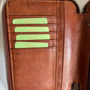 Puede incluir: Una cartera de cuero marr&oacute;n con cierre de cremallera, que presenta cuatro ranuras horizontales para tarjetas en el lado izquierdo. Cada ranura contiene una tarjeta de color verde claro con el texto "VALENGA" impreso. El interior muestra costuras.