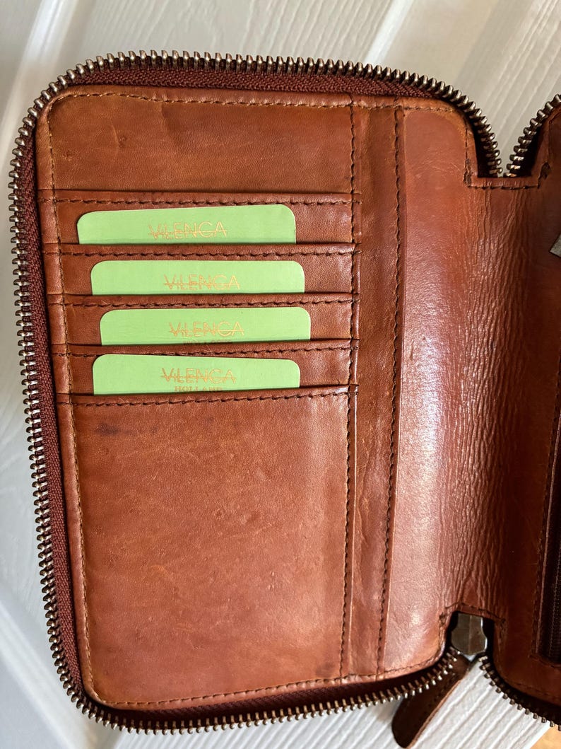 Puede incluir: Una cartera de cuero marr&oacute;n con cierre de cremallera, que presenta cuatro ranuras horizontales para tarjetas en el lado izquierdo. Cada ranura contiene una tarjeta de color verde claro con el texto "VALENGA" impreso. El interior muestra costuras.