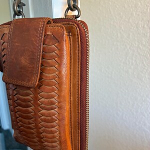 Puede incluir: Un bolso bandolera de cuero color canela con un panel frontal trenzado, cierre de solapa con broche magn&eacute;tico y cremallera lateral. El bolso est&aacute; sujeto a una correa met&aacute;lica mediante dos mosquetones giratorios en la parte superior.