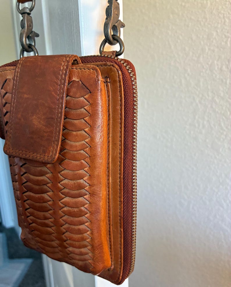 Puede incluir: Un bolso bandolera de cuero color canela con un panel frontal trenzado, cierre de solapa con broche magn&eacute;tico y cremallera lateral. El bolso est&aacute; sujeto a una correa met&aacute;lica mediante dos mosquetones giratorios en la parte superior.