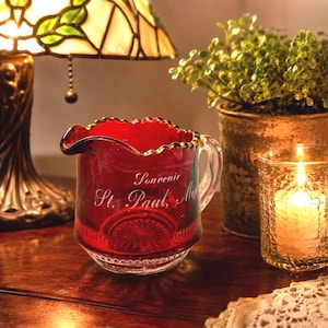 May include: A small, deep red glass creamer pitcher with a scalloped rim and a clear glass handle. The front of the pitcher features etched white text that reads Souvenir St. Paul, Minn. The glass has a textured, pressed pattern at the base.