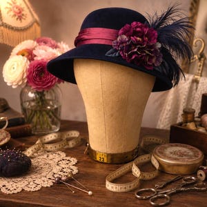 May include: A navy blue felt cloche hat featuring a deep magenta satin ribbon band, a cluster of dark purple fabric flowers, and dark blue feathers. The hat is displayed on a beige mannequin head with a visible metal base marked with the size 22.