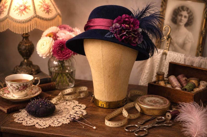 May include: A navy blue felt cloche hat featuring a deep magenta satin ribbon band, a cluster of dark purple fabric flowers, and dark blue feathers. The hat is displayed on a beige mannequin head with a visible metal base marked with the size 22.