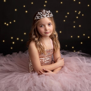 Könnte beinhalten: Ein junges Mädchen in einem rosa Tüllkleid und einer glitzernden Tiara, das vor einem schwarzen Hintergrund mit goldenen Glitzersternen sitzt.