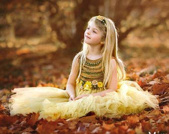 May include: A young girl wearing a yellow tulle dress with a green and gold floral bodice sits in a pile of autumn leaves. She is looking up and smiling.