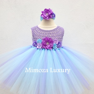 Pu&ograve; includere: Un abito tutu di tulle blu chiaro e viola con un corpetto all'uncinetto e una fascia per capelli con fiori. Il corpetto &egrave; decorato con fiori viola e blu. L'abito ha il testo "Mimosa Luxury" sulla gonna.