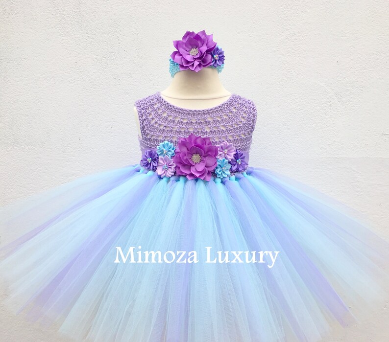 Pu&ograve; includere: Un abito tutu di tulle blu chiaro e viola con un corpetto all'uncinetto e una fascia per capelli con fiori. Il corpetto &egrave; decorato con fiori viola e blu. L'abito ha il testo "Mimosa Luxury" sulla gonna.
