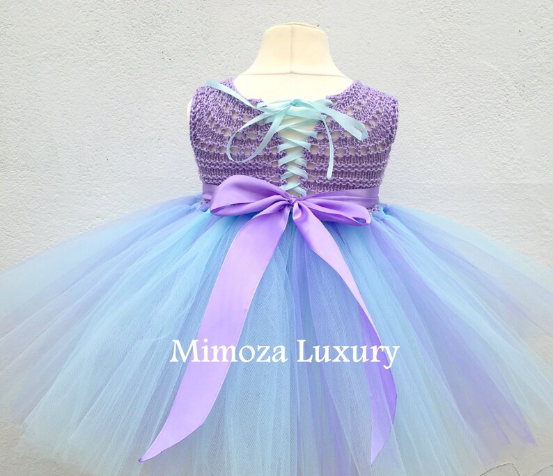 Pu&ograve; includere: Un abito tutu in tulle blu chiaro e viola con un corpetto in crochet lavanda e un nastro di raso lavanda abbinato legato in un fiocco sul retro. Il corpetto ha una schiena con lacci.