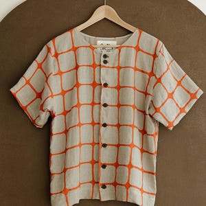 May include: A short-sleeved button-down shirt in a light beige color featuring a repeating grid pattern of bold orange rounded squares. The shirt has a round neckline and a vertical row of dark brown buttons down the center front.