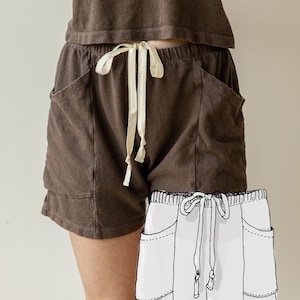 May include: Dark brown shorts featuring an elastic waistband with a cream-colored drawstring, two large side pockets, and a matching brown top. A white line drawing illustration of the shorts is overlaid on the bottom right corner of the image.