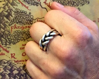 May include: A silver ring with a braided design. The ring is worn on the index finger of a hand.