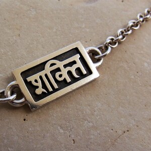 May include: Silver chain necklace with a rectangular pendant featuring the word "शक्ति" in Hindi script.
