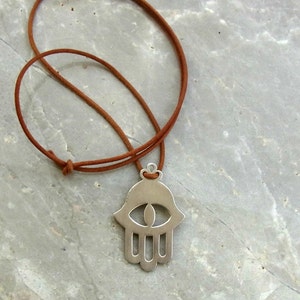 May include: A silver hamsa hand charm necklace with an eye in the center, hanging from a brown leather cord.