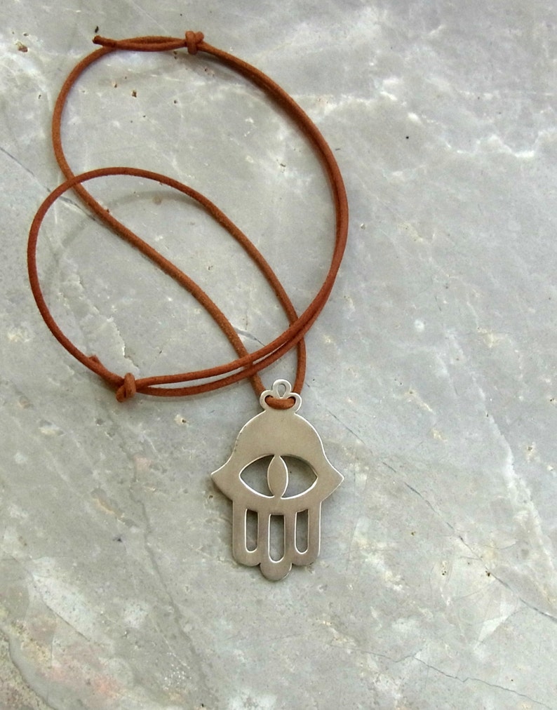May include: A silver hamsa hand charm necklace with an eye in the center, hanging from a brown leather cord.