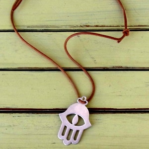 May include: A silver hamsa hand charm necklace with a brown leather cord. The hamsa hand is a symbol of protection and good luck.
