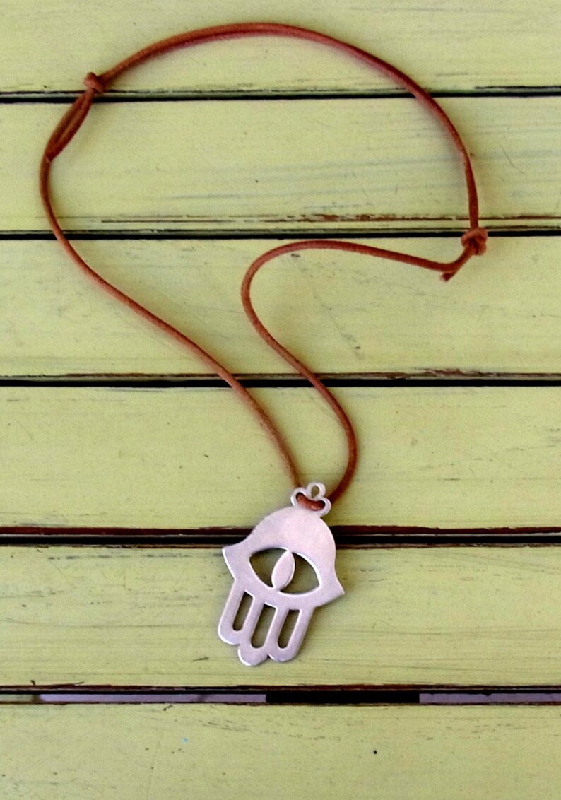 May include: A silver hamsa hand charm necklace with a brown leather cord. The hamsa hand is a symbol of protection and good luck.