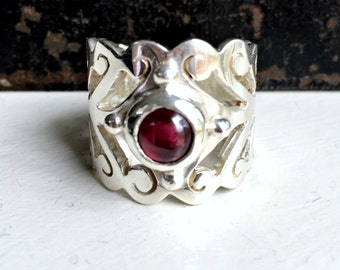 May include: A silver ring with an intricate design and a red gemstone in the center.
