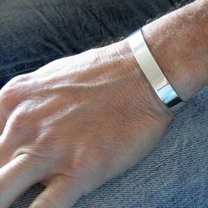 May include: A silver cuff bracelet worn on a person's wrist. The bracelet is wide and flat, with a smooth, polished finish.