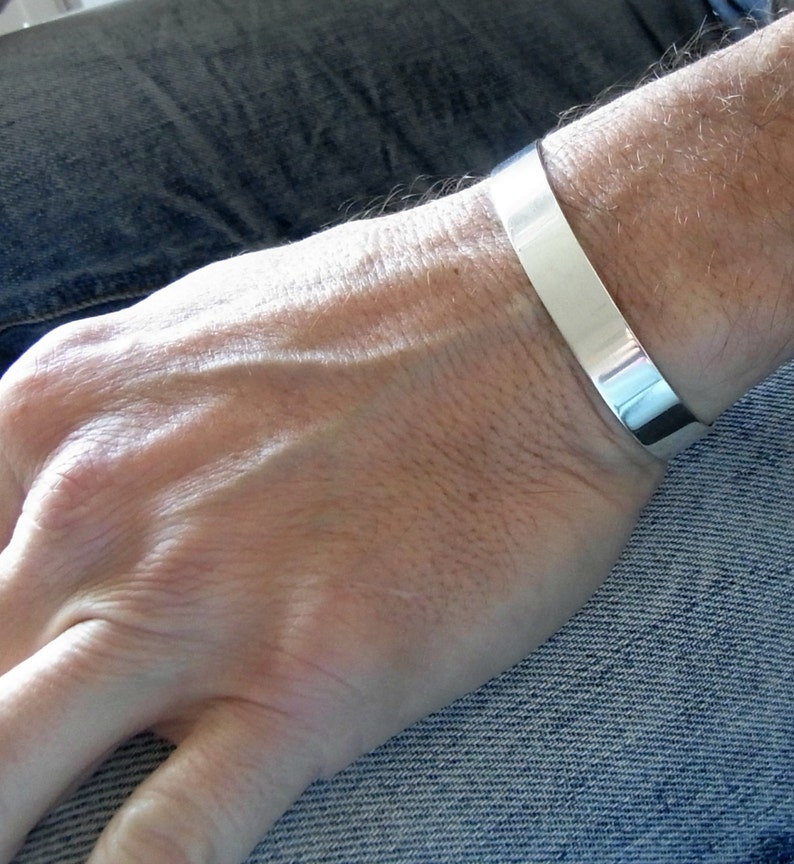 May include: A silver cuff bracelet worn on a person's wrist. The bracelet is wide and flat, with a smooth, polished finish.