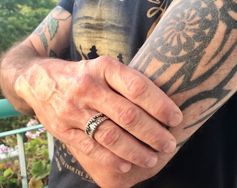 Könnte beinhalten: Ein silberner Ring mit einem strukturierten Design. Der Ring wird am Mittelfinger einer Hand getragen.