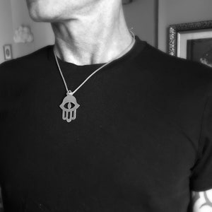 May include: A silver hamsa hand pendant necklace with an eye in the center. The necklace is on a person wearing a black shirt.