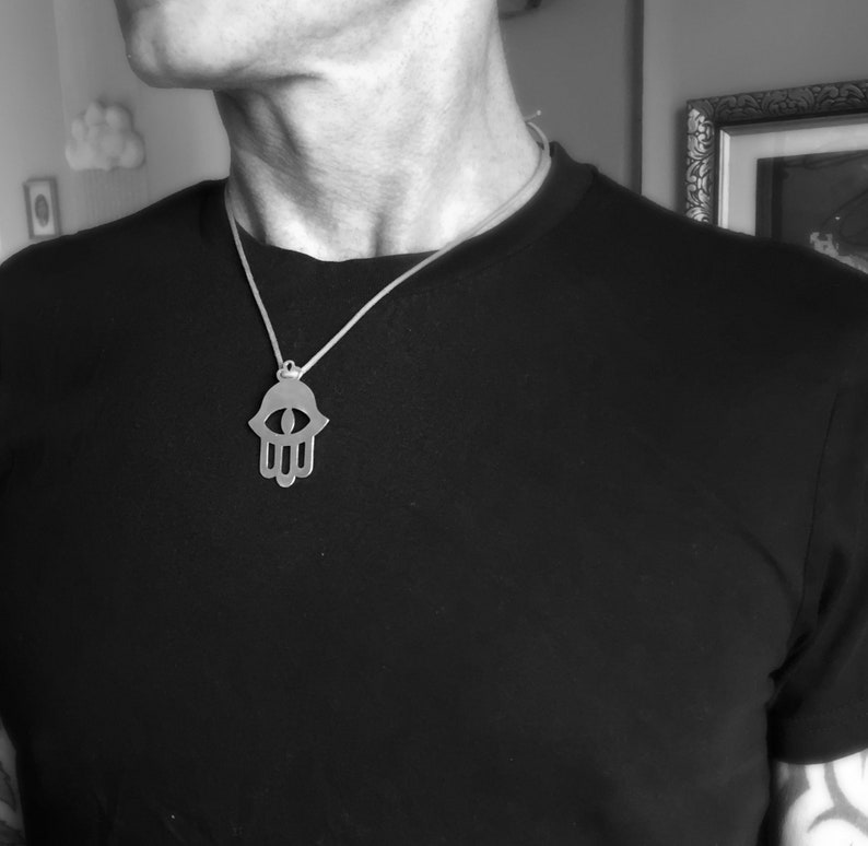 May include: A silver hamsa hand pendant necklace with an eye in the center. The necklace is on a person wearing a black shirt.