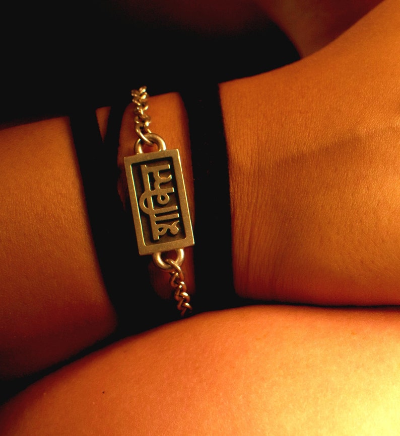 May include: A silver bracelet with a rectangular charm that says "Fuelia" in a stylized font. The bracelet is worn on a person's wrist.