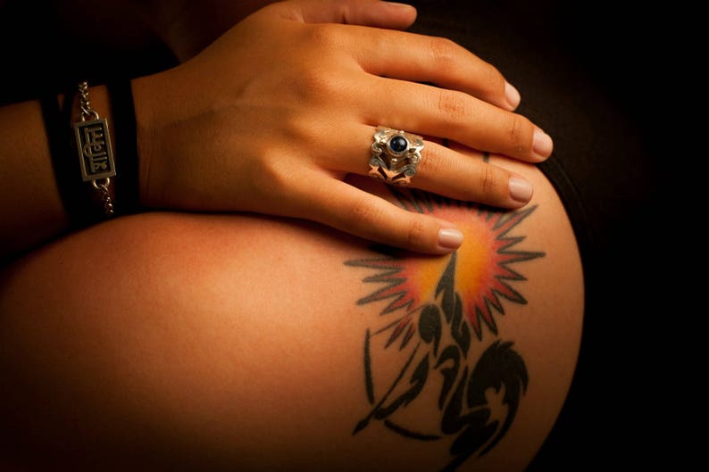May include: A close-up of a person's arm with a silver bracelet that says "Ellie" and a silver ring with a blue stone. The arm has a tattoo of a stylized sun with a black and white design.