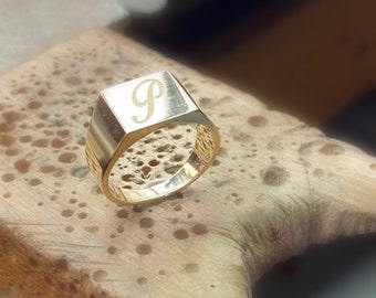 May include: A gold signet ring with a square face, engraved with the letter "P". The ring has a polished finish on the face and a textured band. The ring is a classic piece of jewelry.