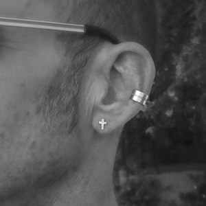 May include: A close-up of a person's ear with a small silver cross earring and a silver ear cuff. The earring is in the lobe and the cuff is on the upper part of the ear.