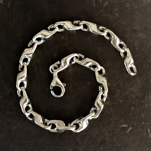 K&ouml;nnte beinhalten: Ein silbernes Kettenarmband mit einem Karabinerverschluss. Die Kette besteht aus ineinandergreifenden Gliedern, die wellenf&ouml;rmig sind.
