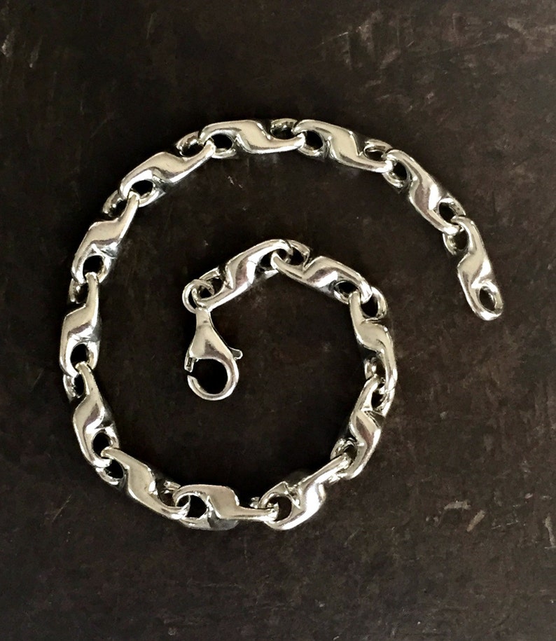 K&ouml;nnte beinhalten: Ein silbernes Kettenarmband mit einem Karabinerverschluss. Die Kette besteht aus ineinandergreifenden Gliedern, die wellenf&ouml;rmig sind.
