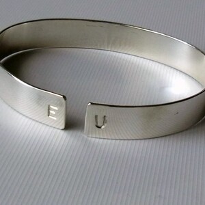 May include: A silver bangle bracelet with a split design, featuring the letters "E" and "U" engraved on each side.