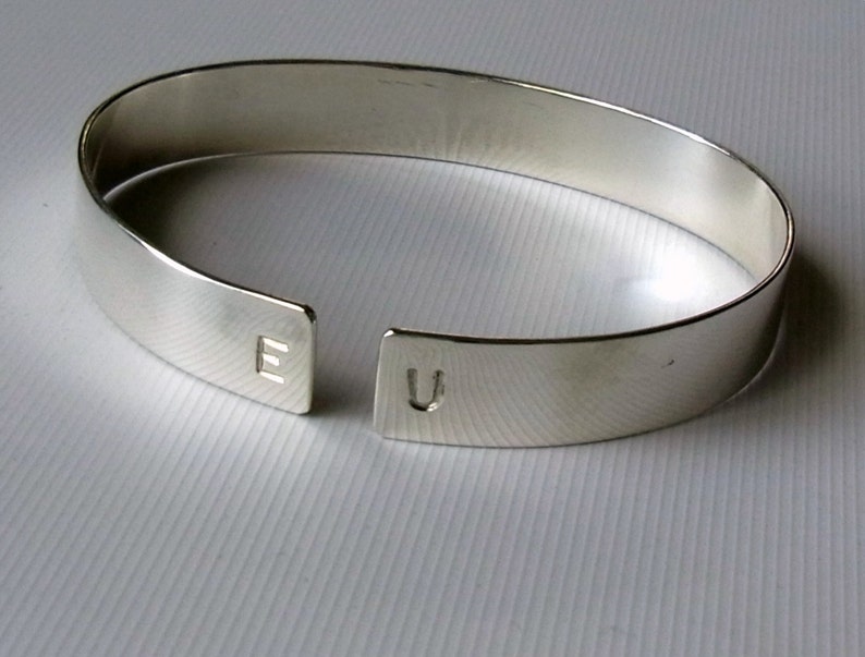 May include: A silver bangle bracelet with a split design, featuring the letters "E" and "U" engraved on each side.