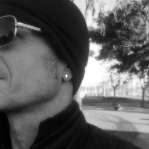 May include: A black and white photo of a person wearing a black beanie and sunglasses. The person is wearing a small cross earring.