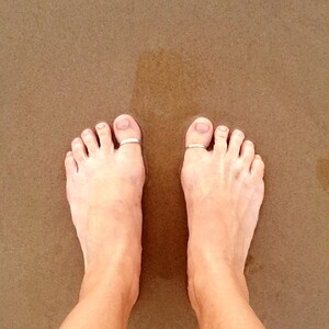 May include: A close-up of two bare feet with silver toe rings on the big toes. The feet are on a sandy surface.