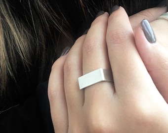 May include: A silver rectangular ring with a smooth, flat surface. The ring is worn on the index finger of a hand.