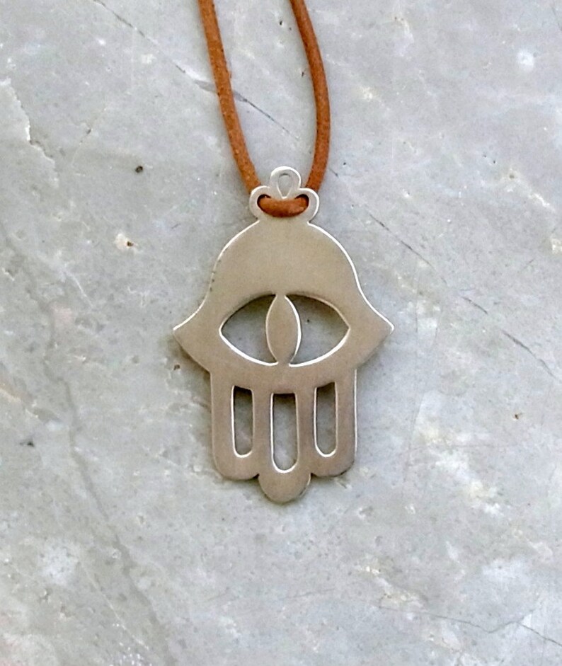 May include: A silver hamsa hand charm necklace with a brown suede cord. The hamsa hand is a symbol of protection and good luck.
