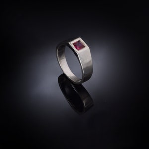 May include: A silver ring with a square, red gemstone set in the center.