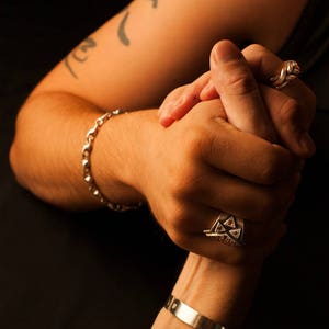 K&ouml;nnte beinhalten: Zwei Personen halten sich an den H&auml;nden, eine Person tr&auml;gt ein silbernes Armband und einen silbernen Ring mit einem dreieckigen Design, die andere Person tr&auml;gt einen silbernen Ring mit einem verdrehten Design.
