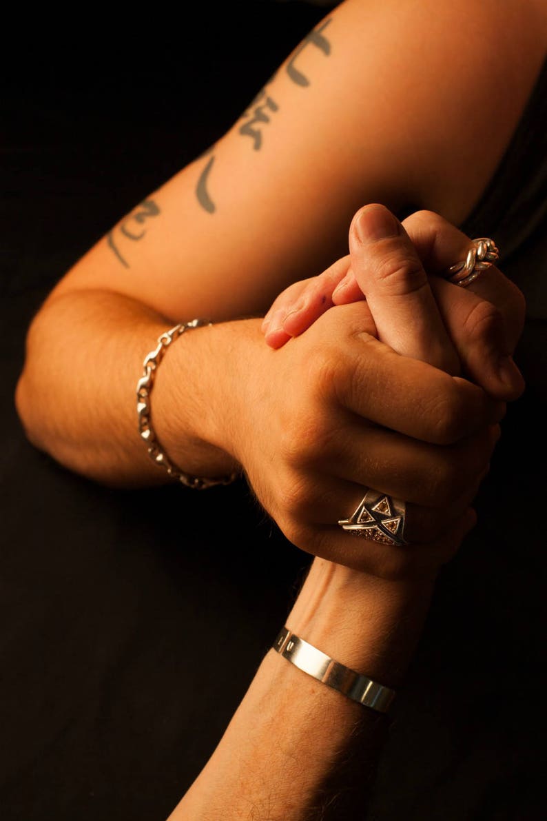 K&ouml;nnte beinhalten: Zwei Personen halten sich an den H&auml;nden, eine Person tr&auml;gt ein silbernes Armband und einen silbernen Ring mit einem dreieckigen Design, die andere Person tr&auml;gt einen silbernen Ring mit einem verdrehten Design.