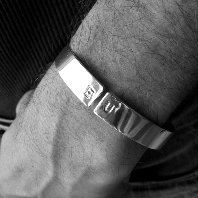 May include: A silver cuff bracelet with a simple design. The bracelet is open on one side and has a hinge on the other. The bracelet is engraved with the letters "M" and "U".