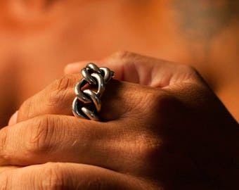 May include: A silver chain link ring with a chunky design. The ring is worn on the index finger of a person's hand.