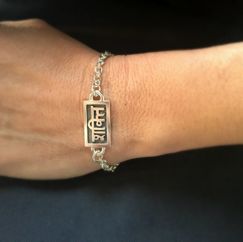 May include: A silver chain bracelet with a rectangular charm that says "शालीन" in black lettering.