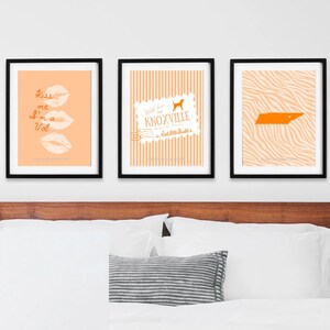 May include: Three framed art prints with black frames. The prints feature orange and white designs, including lips with text, a Knoxville stamp, and a Tennessee outline. The prints are displayed above a bed with white bedding.