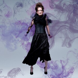 May include: A woman in a black ensemble, including a corset-style top, flowing skirt, and high heels, poses against a backdrop of swirling purple smoke. The outfit features a ruffled collar and wrist cuffs, creating a dramatic, high-fashion look.