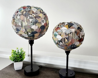 May include: Two table lamps with black bases and round shades made of paper scraps with text from books. The larger lamp has a shade with the text "The Wild Thing Are" and the smaller lamp has a shade with the text "Wild Thing You U".