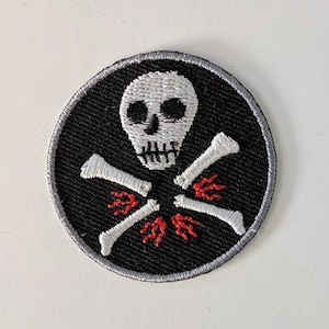 May include: Embroidered patch featuring a skull and crossbones design. The patch is circular, with a black background and a white skull. The crossbones are white, and there are red accents. The patch has a silver border.