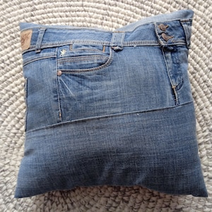 Può includere: Un cuscino in denim blu con una grande tasca sul davanti. Il cuscino è realizzato in denim riciclato e ha una chiusura a bottone.